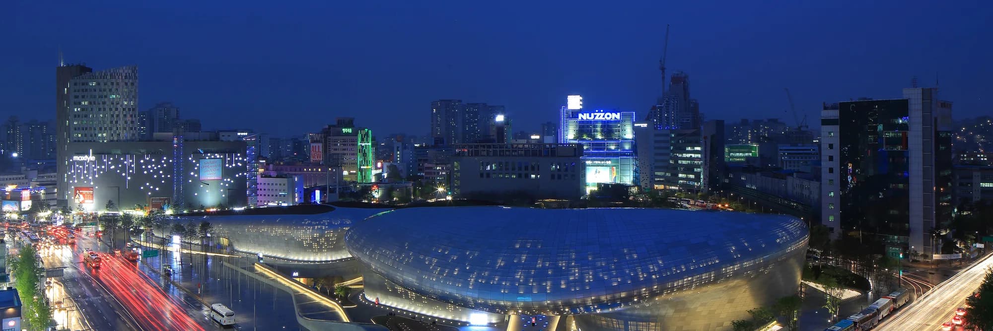 Dongdaemun Design Plaza illuminated at night with the Seoul skyline behind it